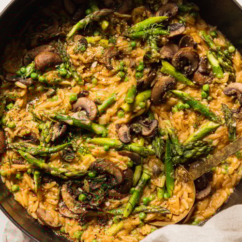 An overhead shot of creamy mushroom asparagus orzo with peas and bits of fresh dill on top. The orzo is in a braiser-style pot with a gold serving utensil sticking out. There is a beige napkin wrapped around the pot handle.