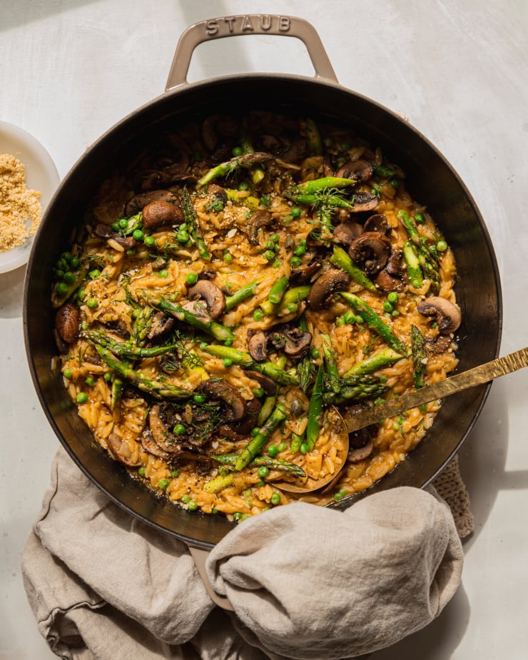 An overhead shot of creamy mushroom asparagus orzo with peas and bits of fresh dill on top. The orzo is in a braiser-style pot with a gold serving utensil sticking out. There is a beige napkin wrapped around the pot handle.