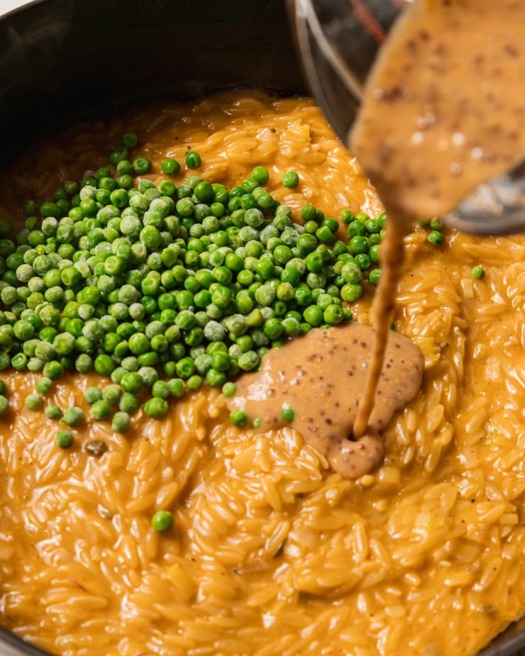 An up close, 3/4 angle image of a pot of creamy orzo with peas on top and a grainy mustard mixture being poured into it.