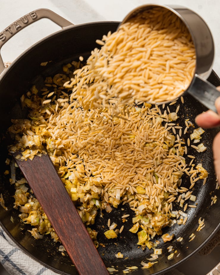 An overhead image shows a hand sprinkling orzo into a pot from a measuring cup.