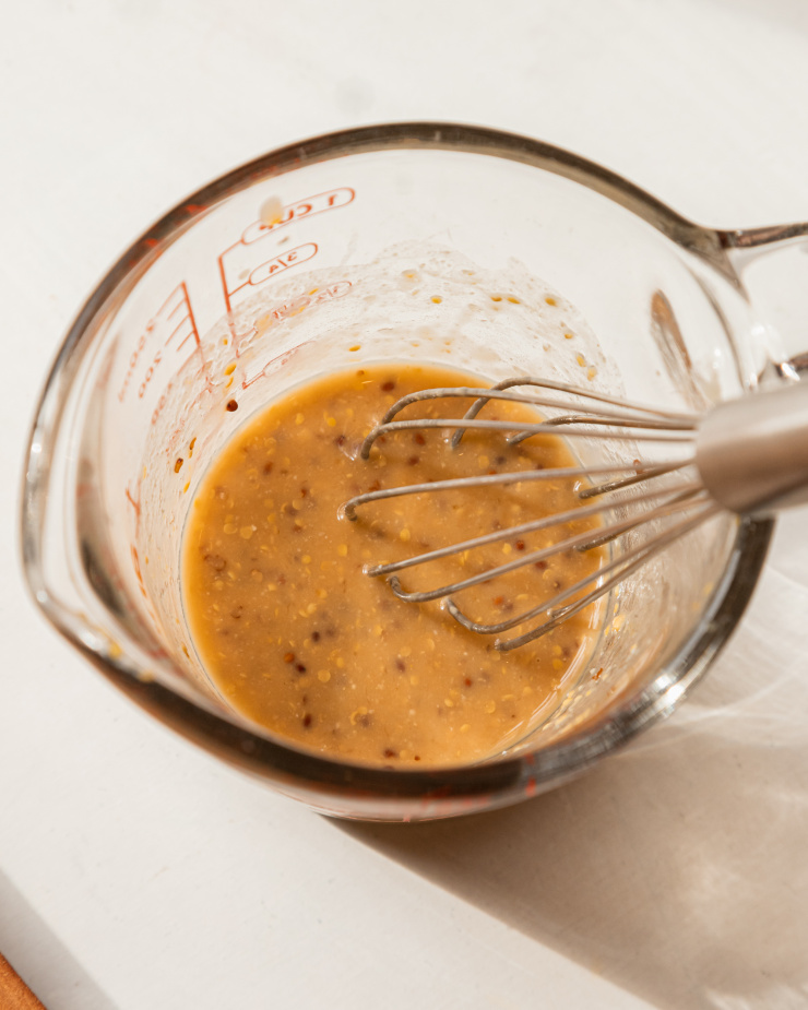 An overhead image shows a grainy mustard, miso, and lemon juice mixture in a glass measuring cup with a whisk sticking out,
