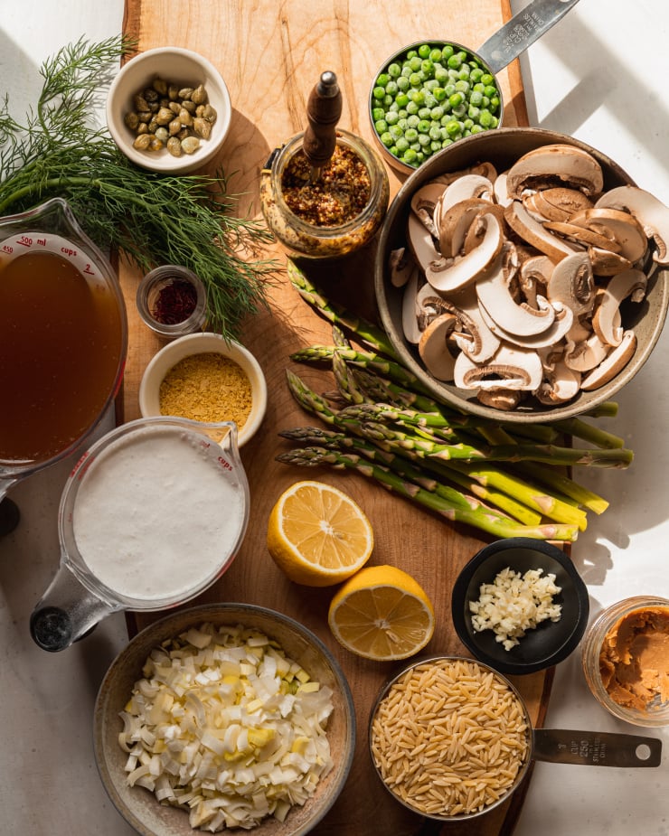 An overhead shot of prepped ingredients for a vegan orzo dish, including a halved lemon, chopped leeks, minced garlic, capers, frozen peas, fresh dill, sliced mushrooms, and spears of asparagus.