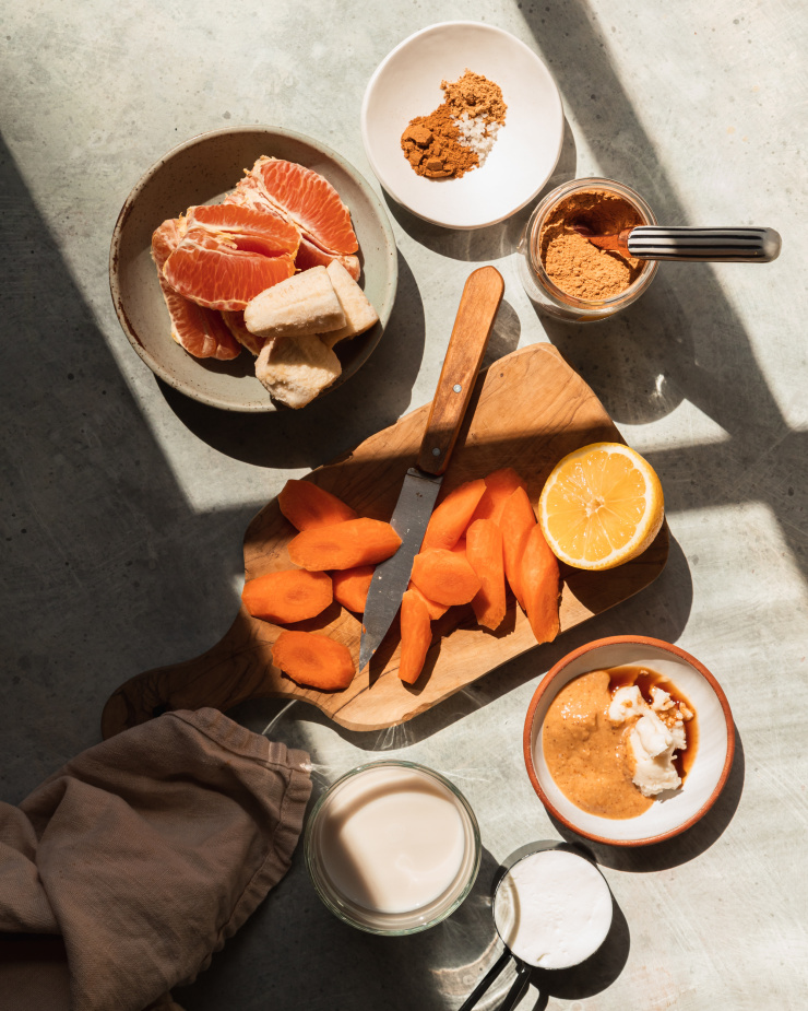 An overhead shot of ingredients for a smoothie, including chopped carrots, a peeled orange, frozen chunks of banana, spices, peanut butter, coconut butter, vegan yogurt, and almond milk.