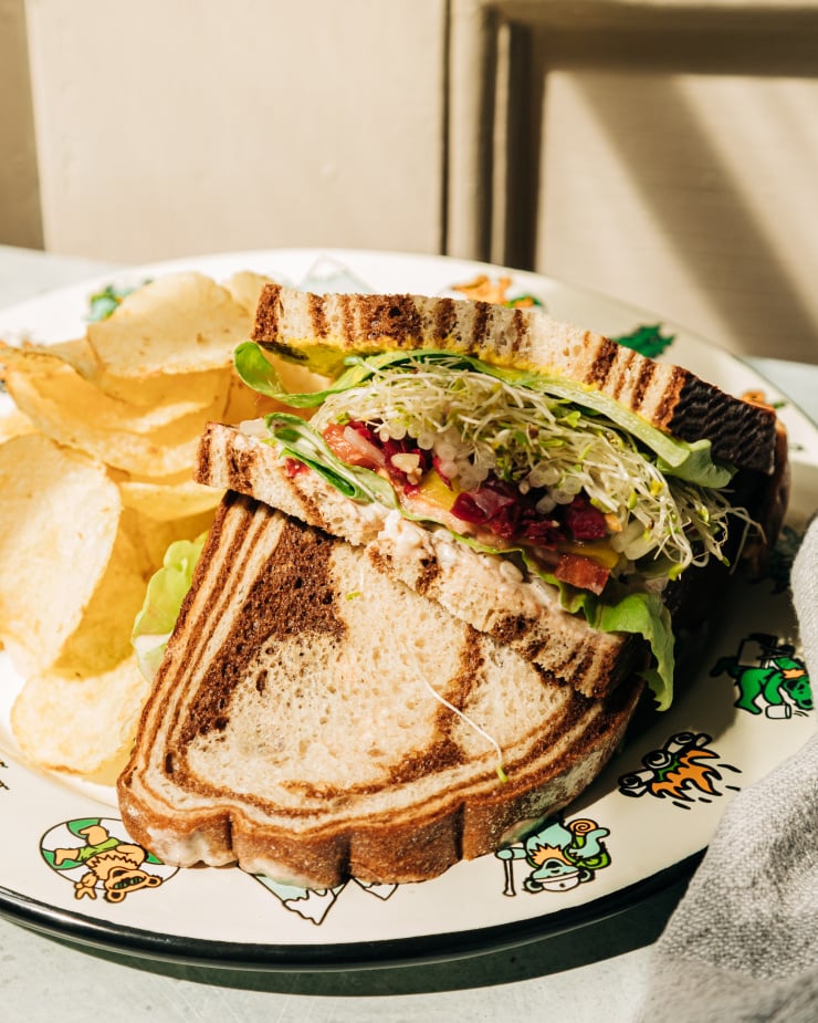 A 3/4 angle shot of a split open veggie crunch sandwich on a plate with some chips. In between marble rye there are layers of sprouts, butter lettuce, a creamy sauce, mustard, tomatoes, sauerkraut, onion, and pepperoncini peppers.
