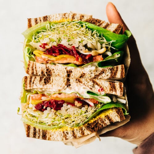 An up close shot of a split open sandwich being held by a hand. In between marble rye there are layers of sprouts, butter lettuce, a creamy sauce, mustard, tomatoes, sauerkraut, onion, and pepperoncini peppers.