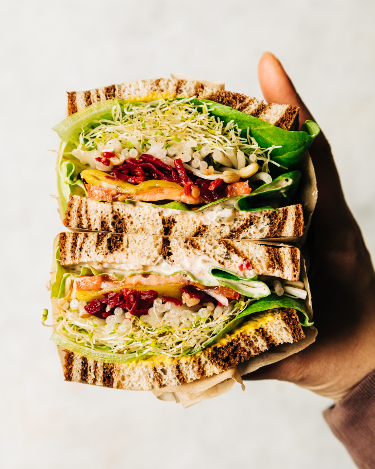 An up close shot of a split open sandwich being held by a hand. In between marble rye there are layers of sprouts, butter lettuce, a creamy sauce, mustard, tomatoes, sauerkraut, onion, and pepperoncini peppers.