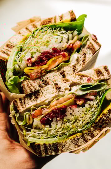 An up close shot of a split open sandwich being held by a hand. In between marble rye there are layers of sprouts, butter lettuce, a creamy sauce, mustard, tomatoes, sauerkraut, onion, and pepperoncini peppers.