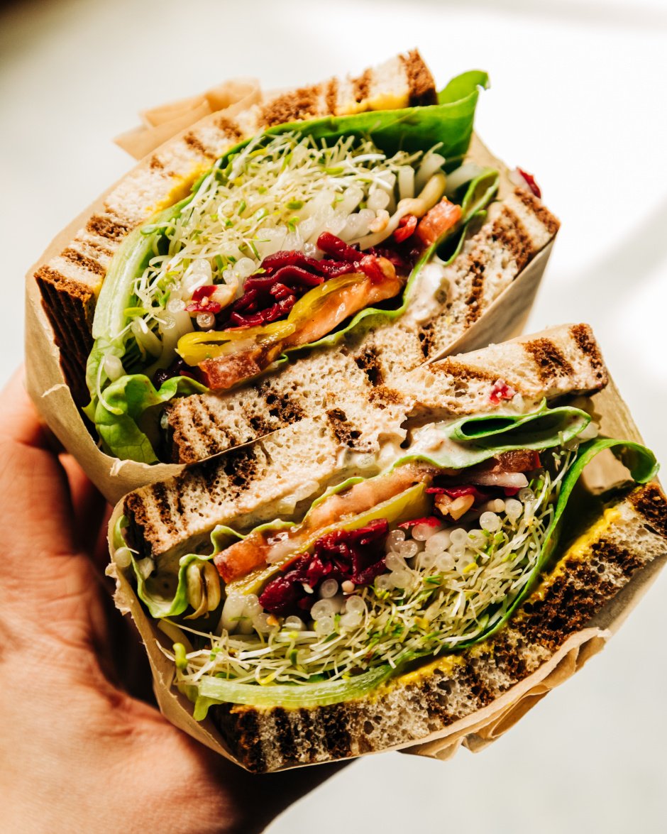 An up close shot of a split open sandwich being held by a hand. In between marble rye there are layers of sprouts, butter lettuce, a creamy sauce, mustard, tomatoes, sauerkraut, onion, and pepperoncini peppers.