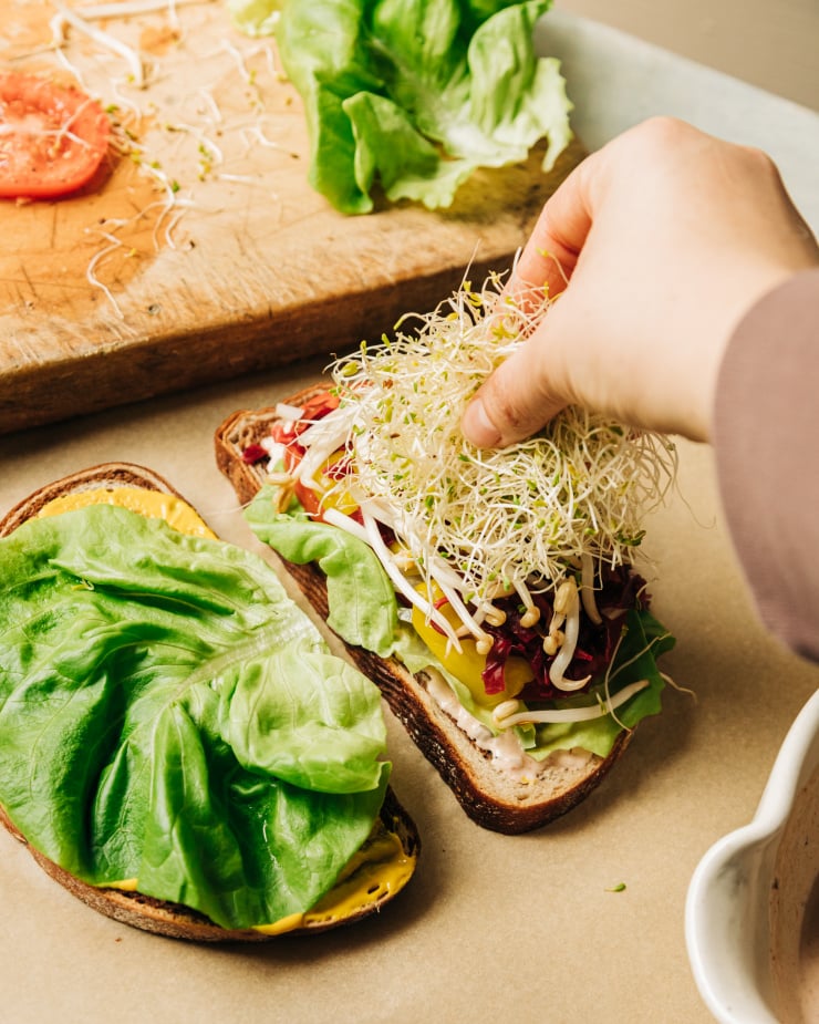 Image shows a hand topping a sandwich with some alfalfa sprouts.