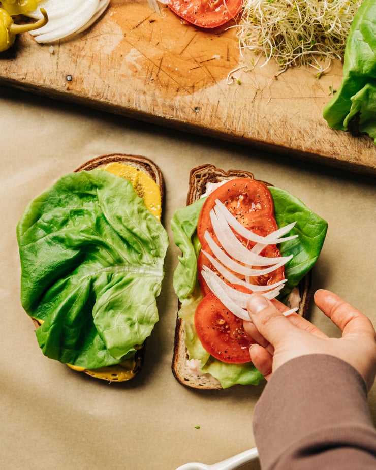 Overhead image shows a hand adding sweet onion slices to a sandwich with lettuce, mustard, and lettuce.
