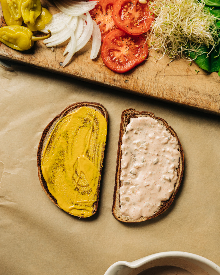 Overhead image shows sandwich components on a wooden board and two pieces of bread topped with mustard and thousand island-style dressing.