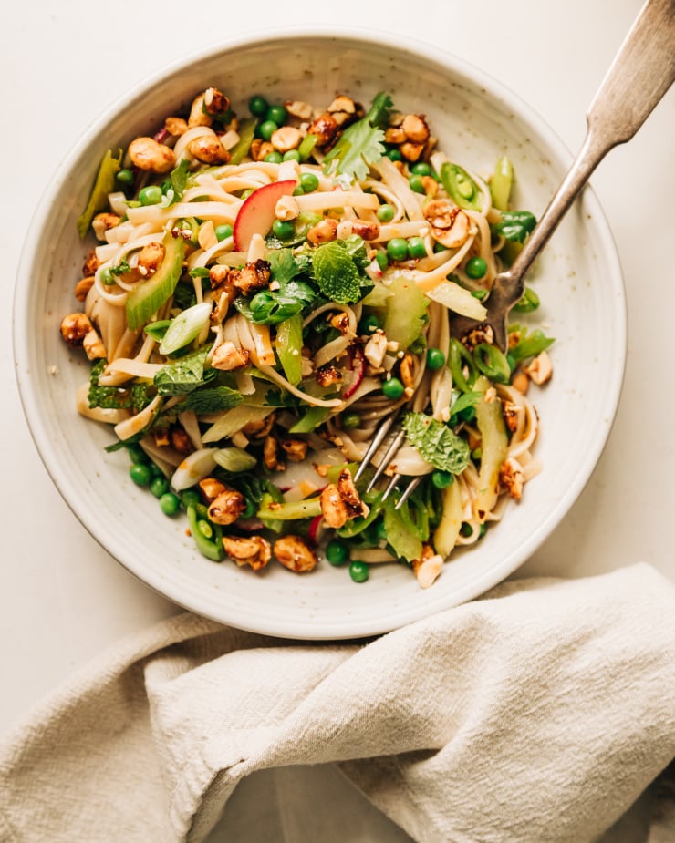 An individual serving of a spring rice noodle salad with peas, snap peas, radishes, sliced celery, mint, cilantro, green onions, and chopped chili maple peanuts on top.