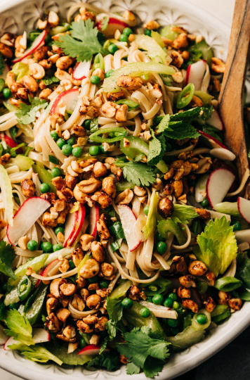 An up close overhead shot of a rice noodle salad in a large bowl with lots of small-diced fresh spring vegetables. The salad is topped with chili and maple syrup roasted peanuts that have been chopped.