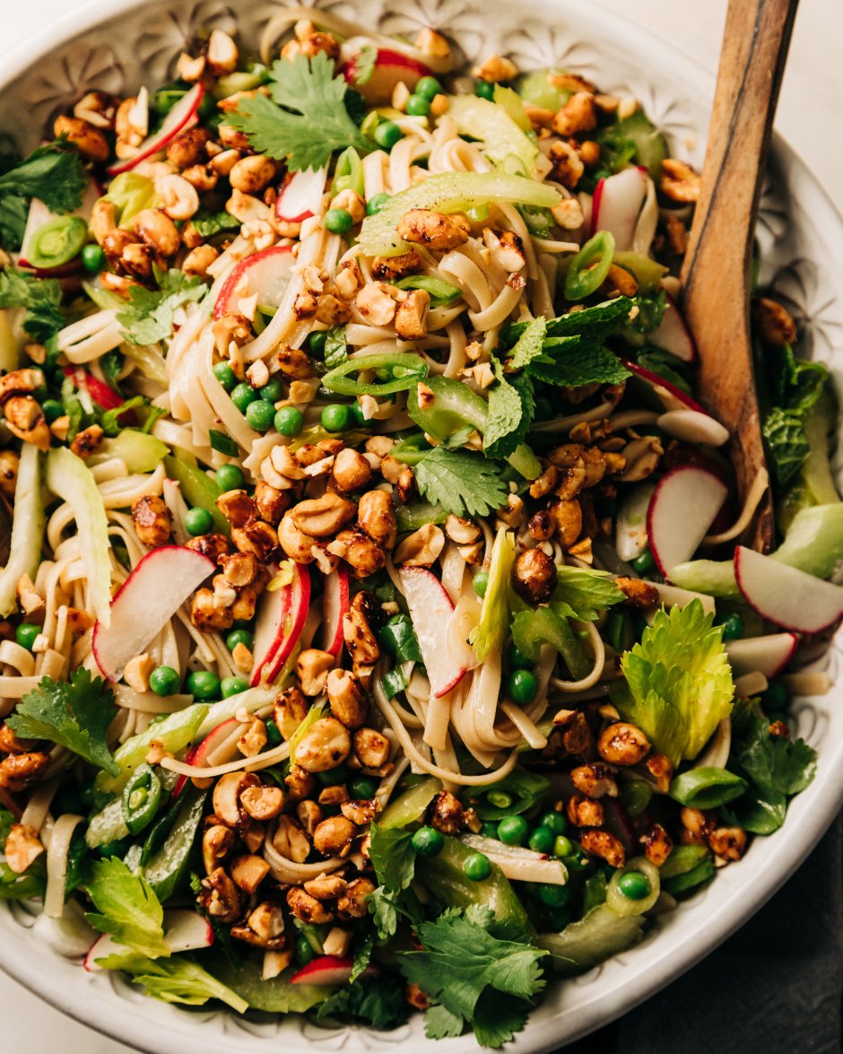 An up close overhead shot of a rice noodle salad in a large bowl with lots of small-diced fresh spring vegetables. The salad is topped with chili and maple syrup roasted peanuts that have been chopped.