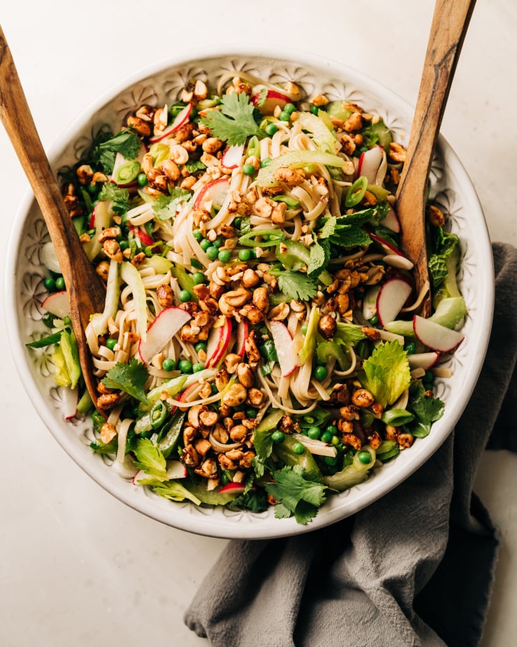 An overhead shot of a rice noodle salad in a large bowl with lots of small-diced fresh spring vegetables. The salad is topped with chili and maple syrup roasted peanuts that have been chopped.