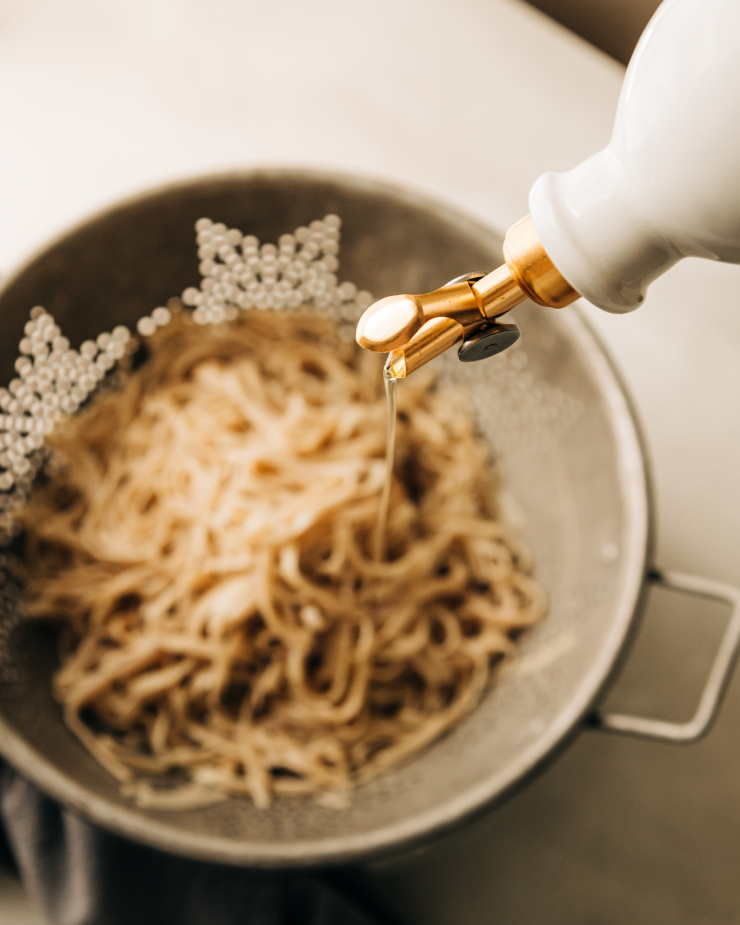 Image shows oil coming from a cream-coloured cruet being poured onto cooked rice noodles in a colander.