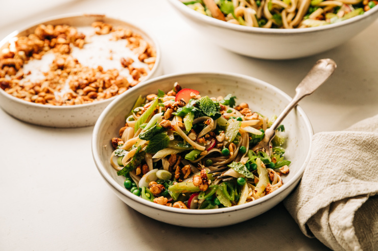 A 3/4 angle shot of an individual serving of a spring rice noodle salad with peas, snap peas, radishes, sliced celery, mint, cilantro, green onions, and chopped chili maple peanuts on top.