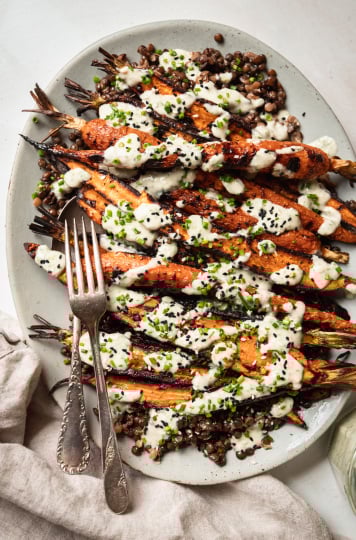 An overhead shot shows a light blue platter topped with cooked french lentils and grilled whole carrots, all topped with a creamy chive-flecked sauce. Extra chives and sesame seeds garnish the dish.