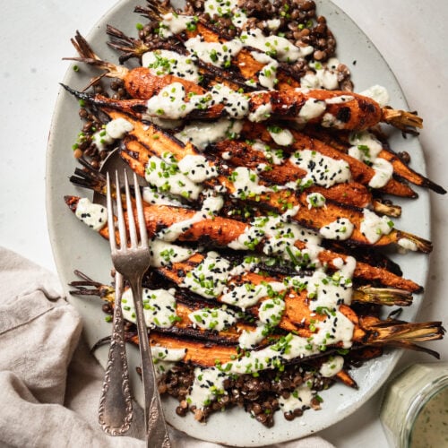 An overhead shot shows a light blue platter topped with cooked french lentils and grilled whole carrots, all topped with a creamy chive-flecked sauce. Extra chives and sesame seeds garnish the dish.