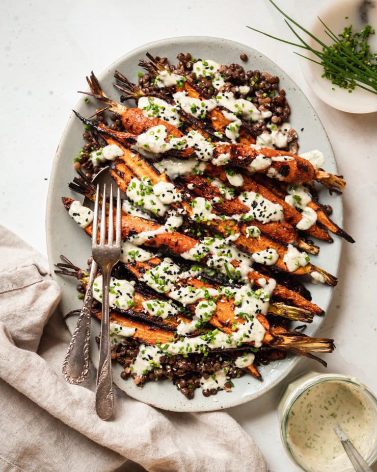 An overhead shot shows a light blue platter topped with cooked french lentils and grilled whole carrots, all topped with a creamy chive-flecked sauce. Extra chives and sesame seeds garnish the dish.