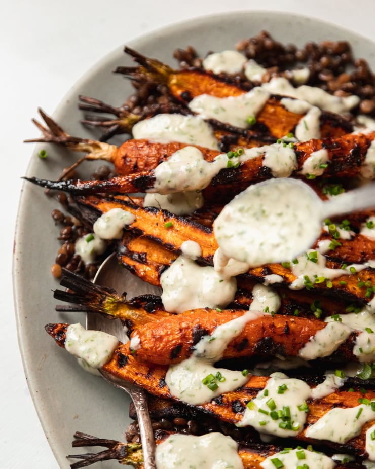 Overhead image shows a spoon drizzling a creamy sauce on top of grilled carrots and lentils, on a pale blue plate.