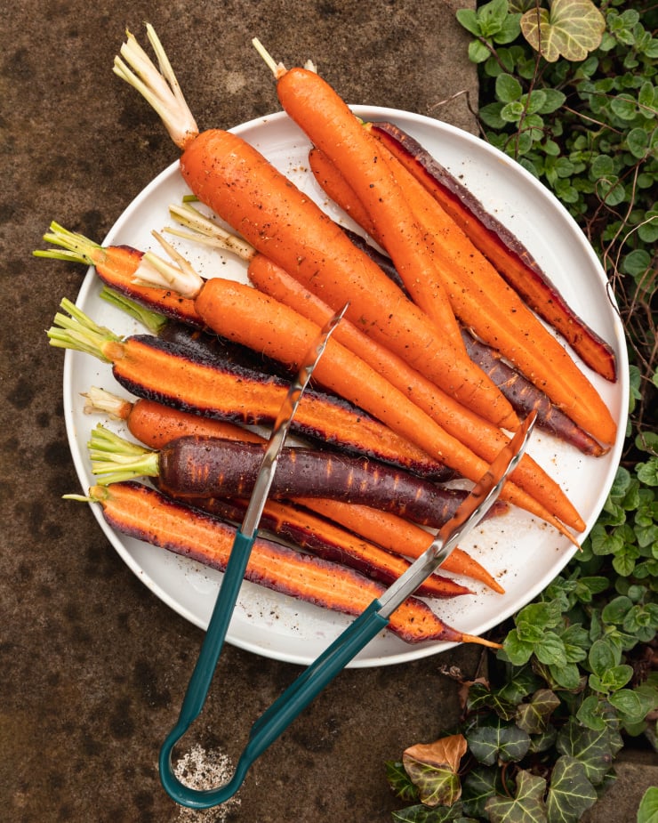 Overhead image shows purple and orange carrots in a white plate with a set of tongs, outdoors.