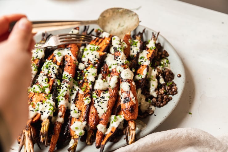 An 3/4 angle shot shows a light blue platter topped with cooked french lentils and grilled whole carrots, all topped with a creamy chive-flecked sauce. Extra chives and sesame seeds garnish the dish. A hand can be seen holding a spoon, drizzling the sauce.