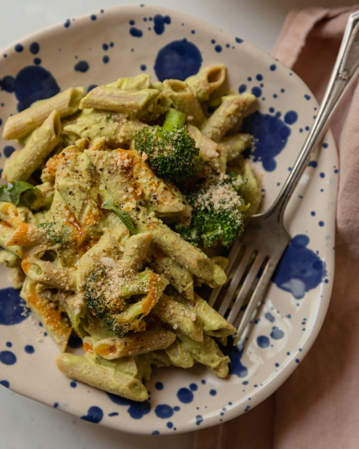 An up close, overhead shot of a single serving of creamy green-sauced pasta that has basil and broccoli in it. The serving bowls are beige and splattered with royal blue dots.