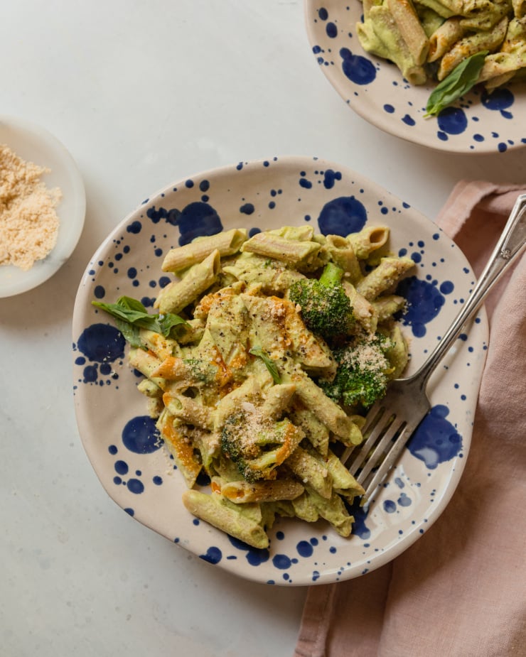 An overhead shot of a single serving of creamy green-sauced pasta that has basil and broccoli in it. The serving bowls are beige and splattered with royal blue dots.