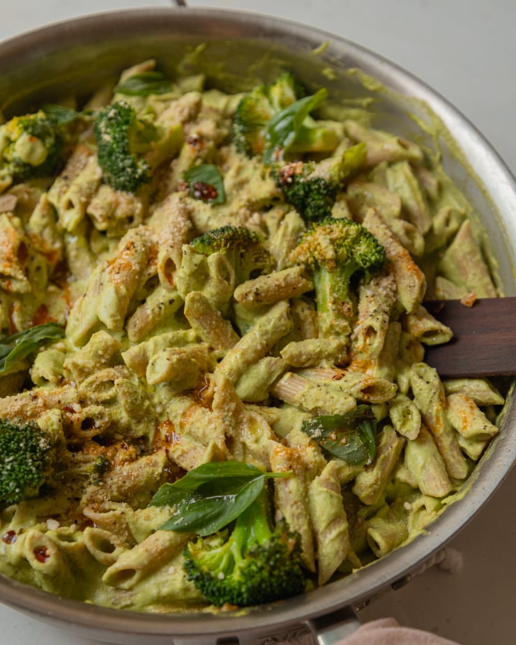 A 3/4 angle shot of a creamy, green-sauced penne pasta topped with cooked broccoli florets and drizzles of deep red chili oil.