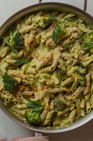 An overhead shot of a creamy green-sauced pasta with broccoli florets and drizzles of deep red chili oil. The 30 minute broccoli basil pasta is shown in a large stainless steel sauté pan.