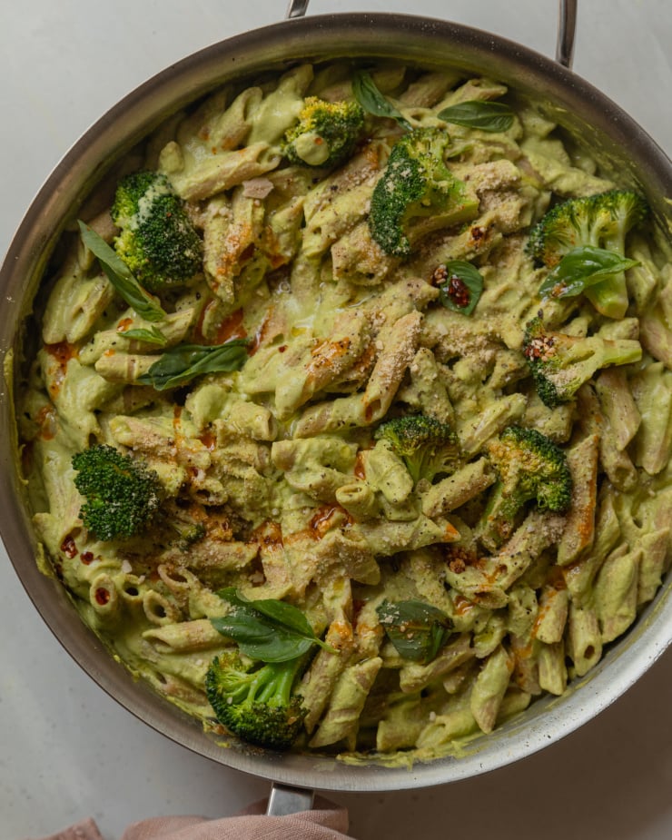 An overhead shot of a creamy green-sauced pasta with broccoli florets and drizzles of deep red chili oil. The 30 minute broccoli basil pasta is shown in a large stainless steel sauté pan.