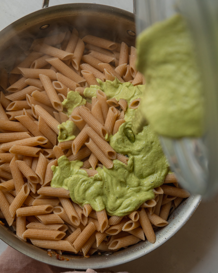 An overhead shot of a creamy green sauce being poured over a pan of cooked whole wheat penne pasta.