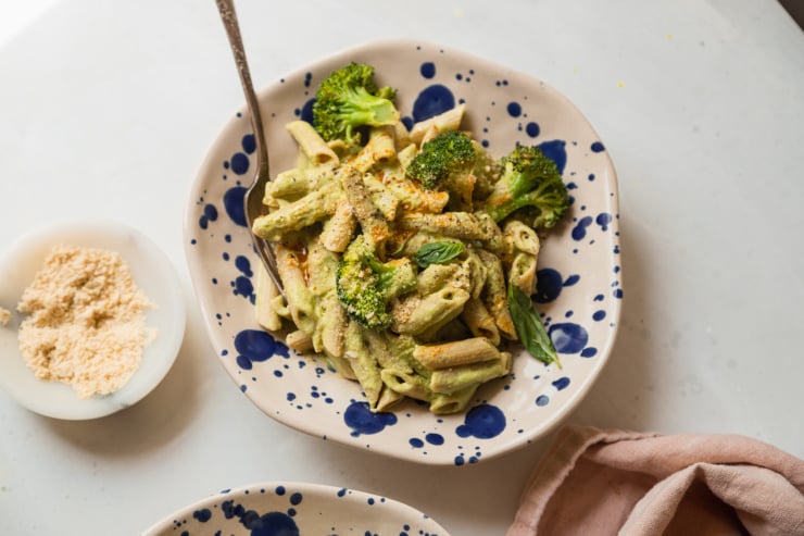 An overhead shot of a single serving of creamy green-sauced pasta that has basil and broccoli in it. The serving bowls are beige and splattered with royal blue dots.