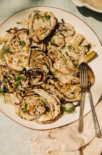 An overhead shot of grilled cabbage and fennel in direct sunlight. The vegetables are covered in a creamy mustard seed sauce and garnished with fennel fronds.