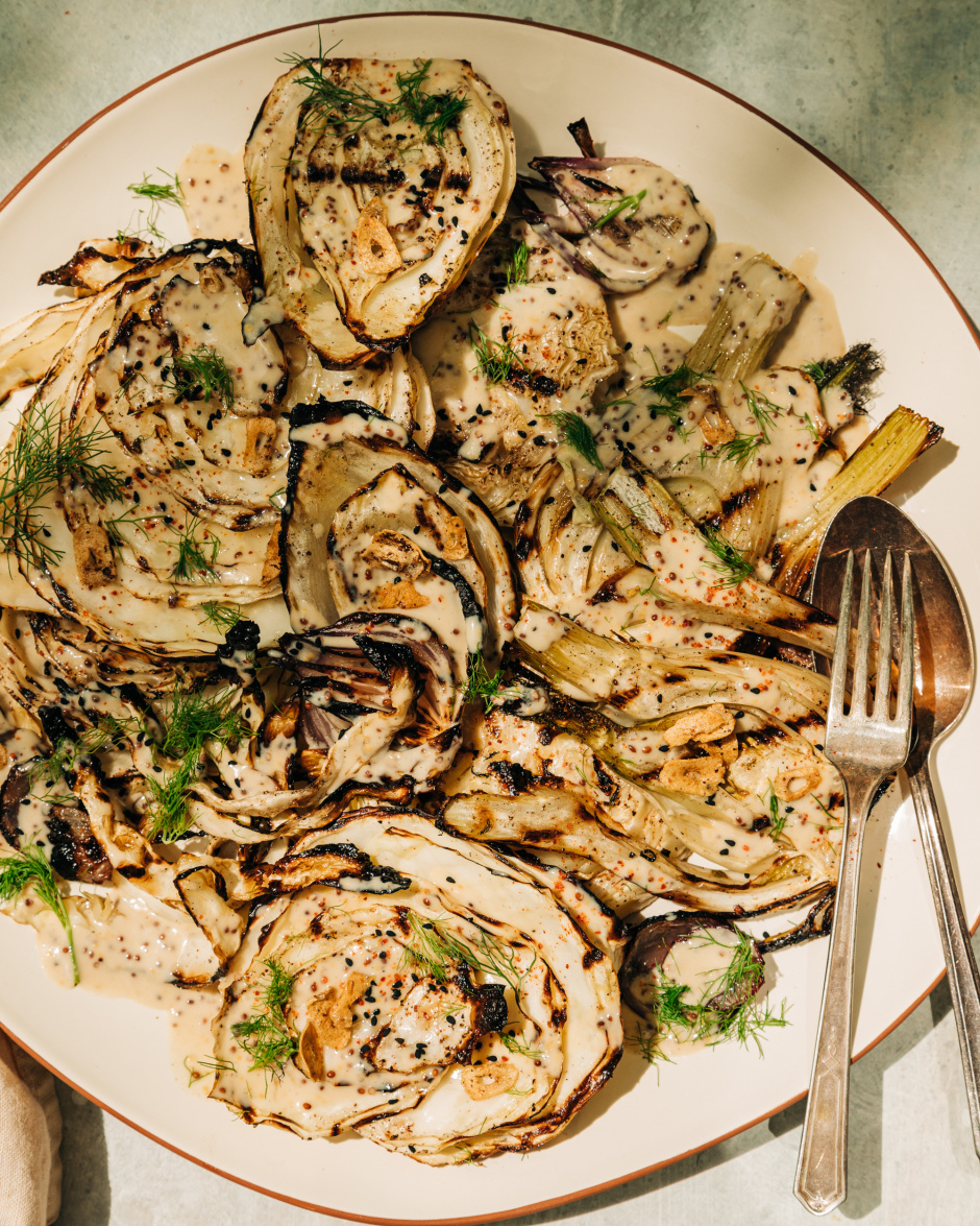 An overhead shot of grilled cabbage and fennel in direct sunlight. The vegetables are covered in a creamy mustard seed sauce and garnished with fennel fronds.
