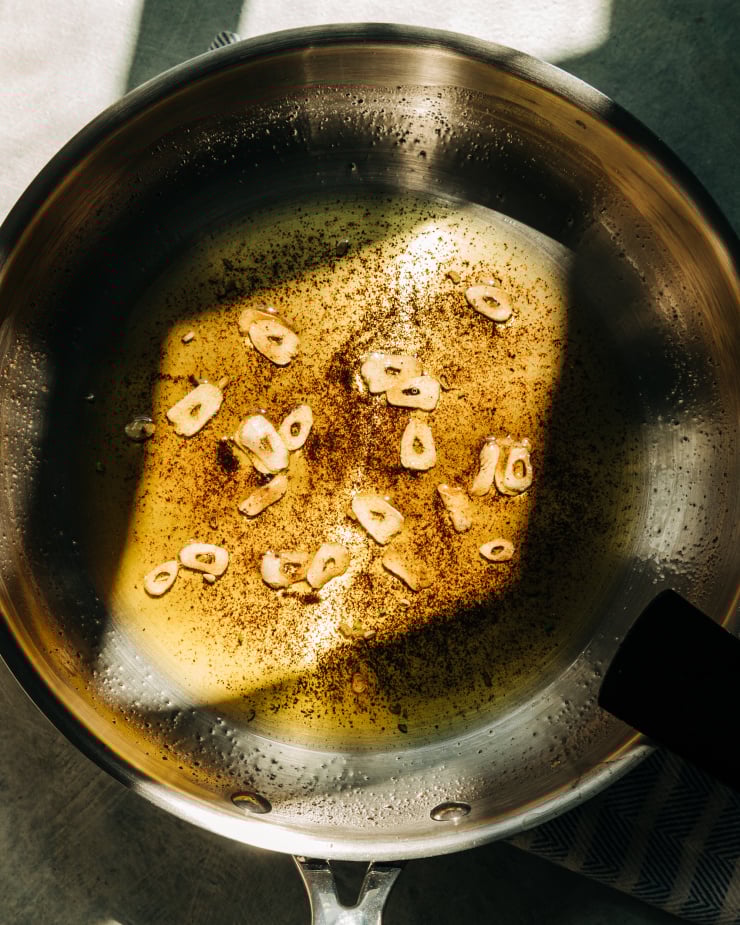 An overhead shot shows a skillet with garlic and sumac infused olive oil in harsh, heavily shadowed light.
