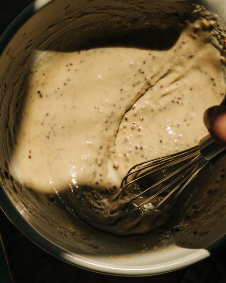 Overhead image shows a hand using a whisk to stir up a vegan maple mustard cream in an off white bowl.