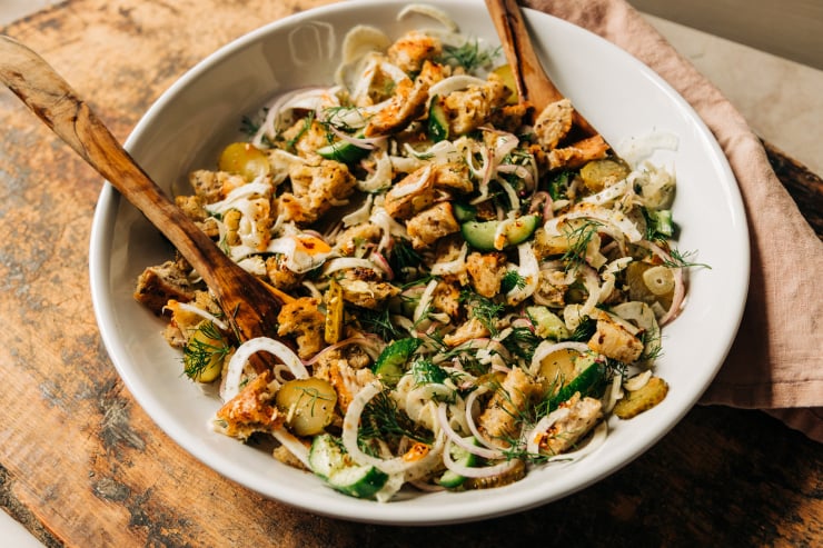 A 3/4 angle shot of a pickle panzanella salad with crispy bits of bread, chopped cucumbers, pickles and red onion, plus shaved fennel and fresh dill. Photographed in a wide, white bowl with wood serving tongs.