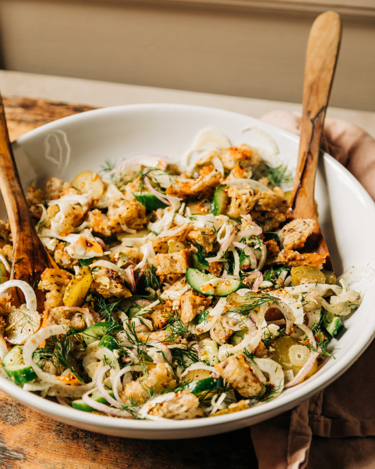 A 3/4 angle shot of a pickle panzanella salad with crispy bits of bread, chopped cucumbers, pickles and red onion, plus shaved fennel and fresh dill. Photographed in a wide, white bowl with wood serving tongs.