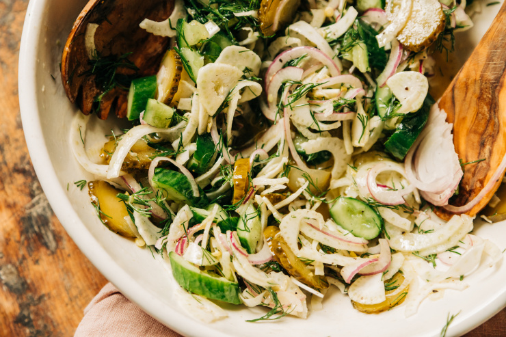 Overhead image shows lightly dressed cucumber, fennel, red onion, pickles, and fresh dill in an off-white bowl with wooden serving tongs.