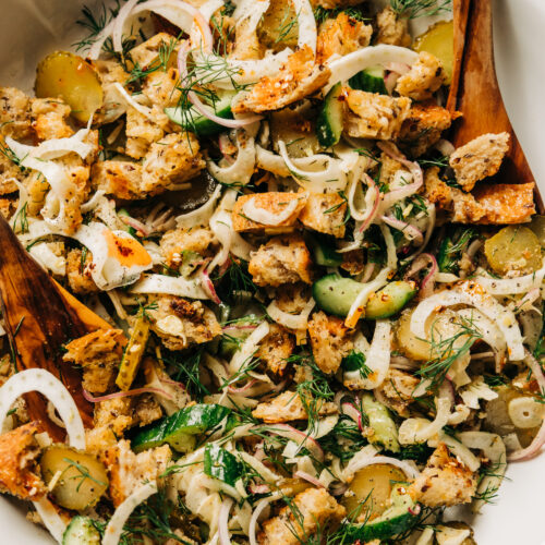 An up close, overhead shot of a pickle panzanella salad with crispy bits of bread, chopped cucumbers, pickles and red onion, plus shaved fennel and fresh dill. Photographed in a wide, white bowl with wood serving tongs.