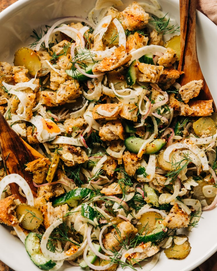 An up close, overhead shot of a pickle panzanella salad with crispy bits of bread, chopped cucumbers, pickles and red onion, plus shaved fennel and fresh dill. Photographed in a wide, white bowl with wood serving tongs.