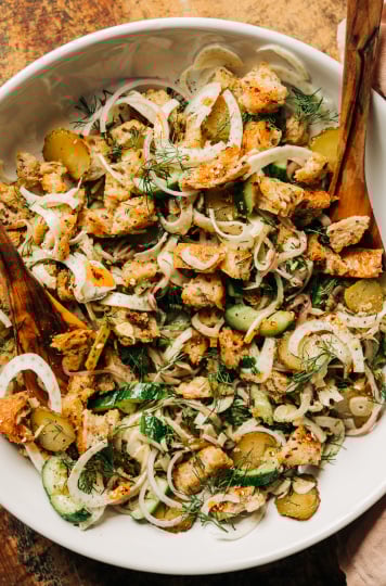 An overhead shot of a pickle panzanella salad with crispy bits of bread, chopped cucumbers, pickles and red onion, plus shaved fennel and fresh dill. Photographed in a wide, white bowl with wood serving tongs. All set on a worn wood background.