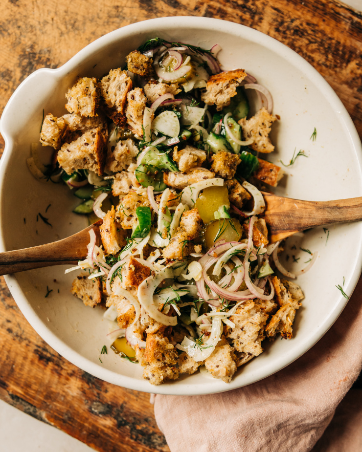 A salad with cucumbers, pickles, fennel, and crispy bits of bread is tossed together in an off-white bowl on a worn wood background.