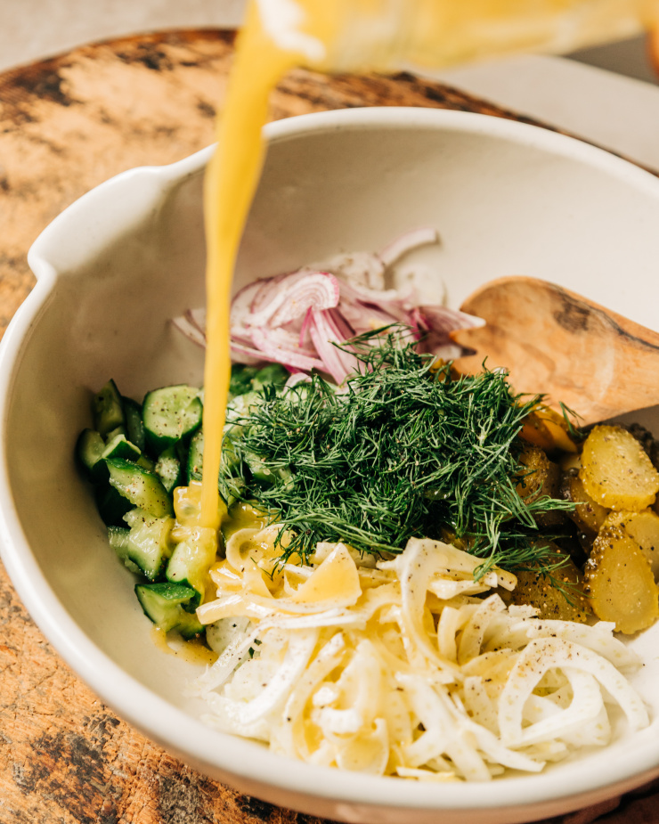 A 3/4 angle image shows a homemade vinaigrette being poured over chopped cucumbers, red onion, fennel, dill, and chopped pickles.