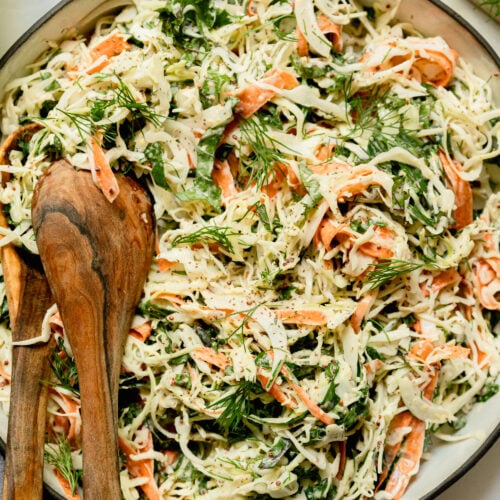 An up close, overhead shot of a cabbage, kale, and carrot slaw with creamy mustard dressing. The slaw is in a wide ceramic dish with a black rim.