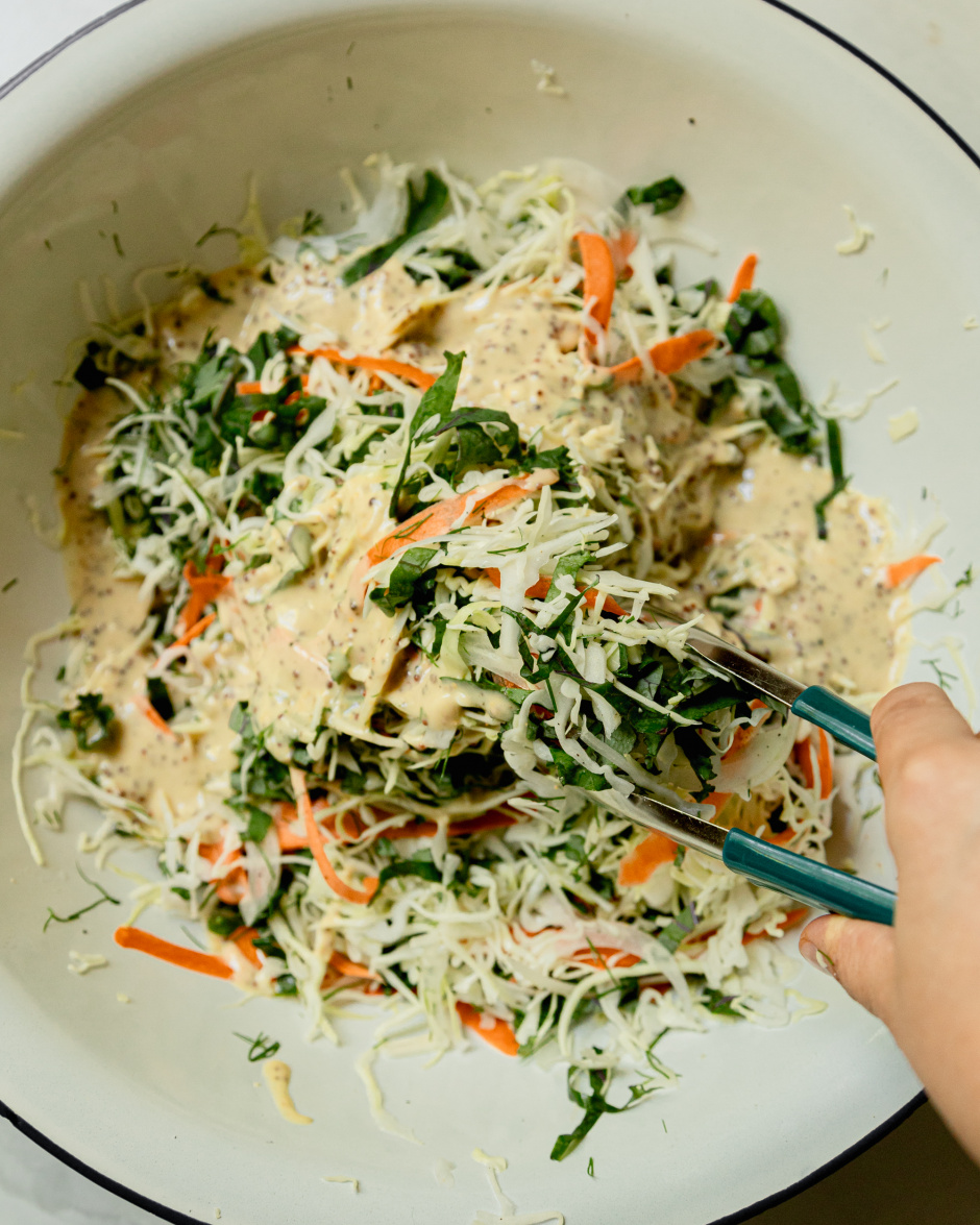 An overhead shot of a hand using some tongs to toss together a creamy mustard slaw in a white bowl.