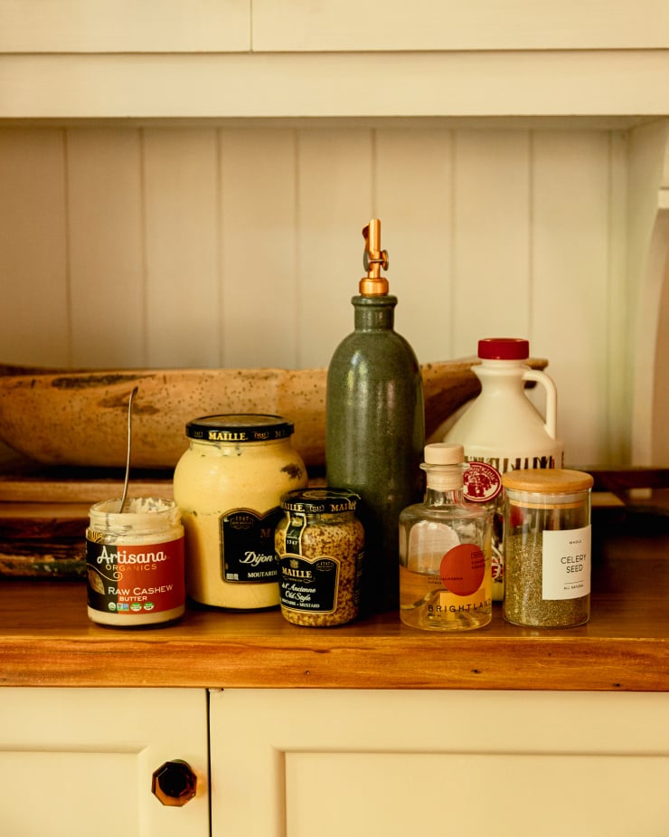 A head on shot of jarred ingredients used for a creamy mustard dressing, all against a white background with some wooden cutting boards and a wooden bread bowl in the background as well.