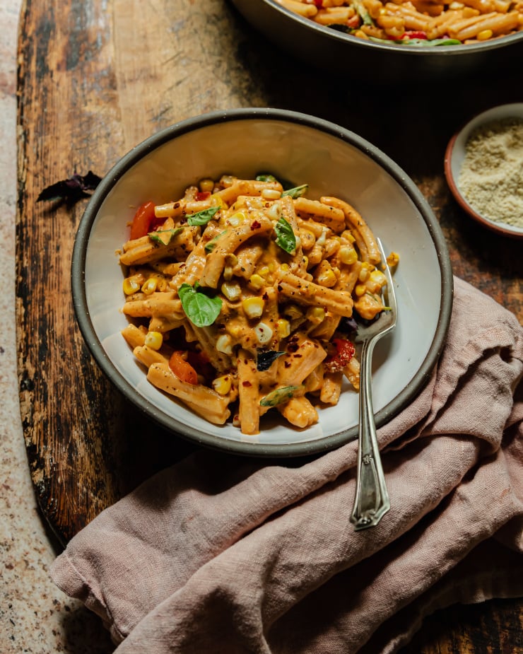 A slight 3/4 angle image shows a bowl of creamy red pepper pasta that features charred corn and strips of roasted red pepper. The pasta is garnished with fresh basil, dried chillies, and ground black pepper. The bowl is seen on a worn wooden board with a vintage silver fork sticking out of the bowl.